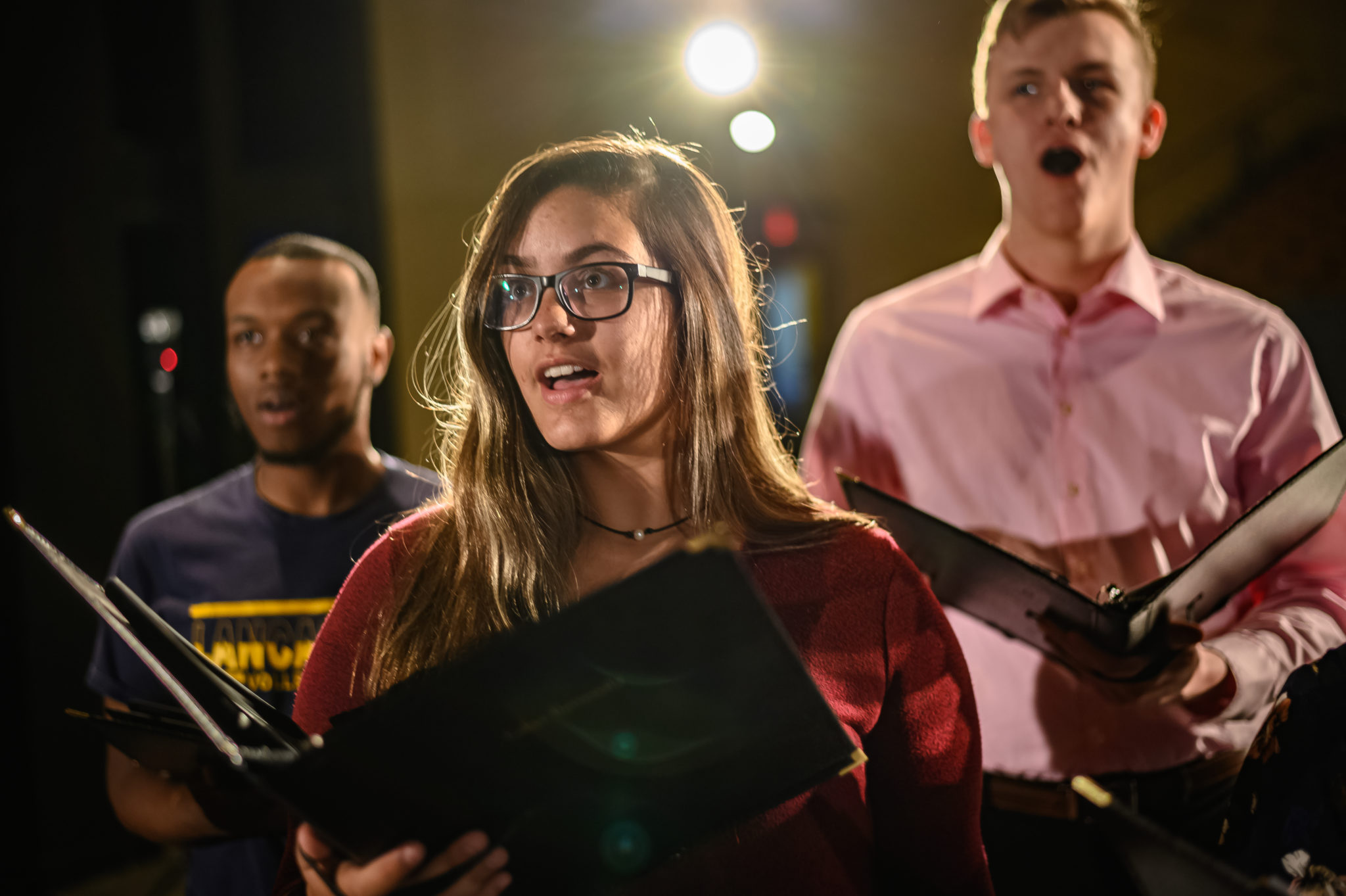 Three singers holding folders, close to one another, on stage