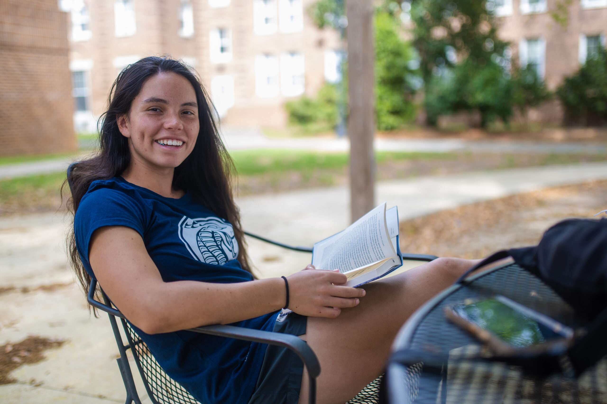 student lounges in a chair with a book in her hand smiling at the camera